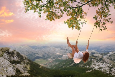 Dream world. Young woman swinging over mountains under sunset sky 
