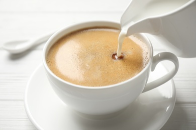Pouring milk into cup of hot coffee on white wooden table, closeup