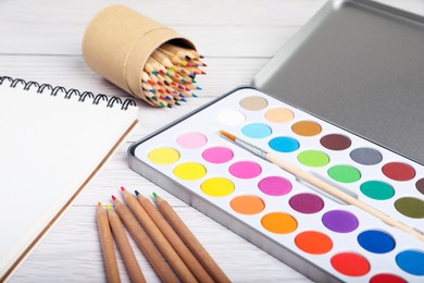 Photo of Watercolor palette with brush, colorful pencils and notebook on white wooden table, closeup