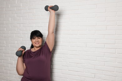 Happy overweight mature woman doing exercise with dumbbells near white brick wall, space for text