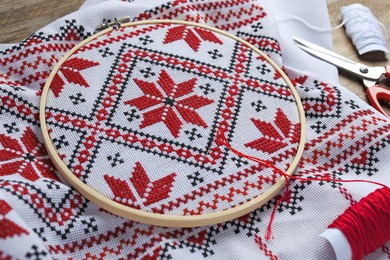 White fabric with red Ukrainian national embroidery in hoop, needle, scissors and threads on table, closeup