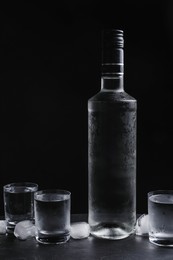 Bottle of vodka and shot glasses with ice on table against black background