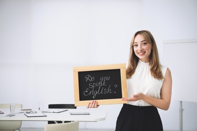 Young female teacher holding chalkboard with words DO YOU SPEAK ENGLISH? in classroom. Space for text