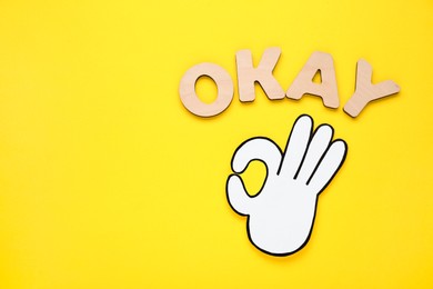 Photo of Word Okay made of wooden letters and paper cutout (OK hand gesture) on yellow background, flat lay. Space for text