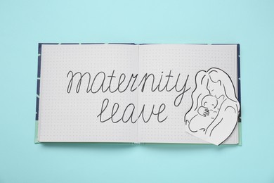 Photo of Notebook with phrase Maternity Leave, paper cutout of mother and child on light blue background, top view