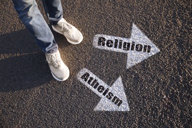 Image of Choice between atheism and religion. Man standing near drawn marks on road, closeup. White arrows pointing in different directions
