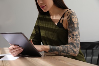 Photo of Beautiful woman with tattoos on arm using tablet at table indoors, closeup