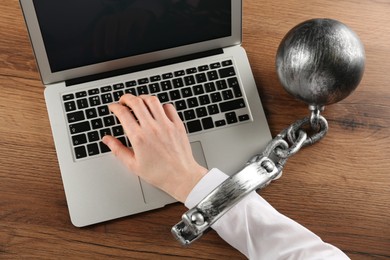 Woman shackled with ball and chain typing on laptop at wooden table, above view. Internet addiction