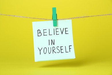 Photo of Card with phrase Believe In Yourself hanging on rope against yellow background. Motivational quote