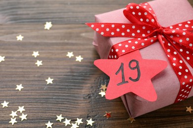 Saint Nicholas Day. Wrapped gift box with date December 19 and star shape confetti on wooden table, closeup. space for text