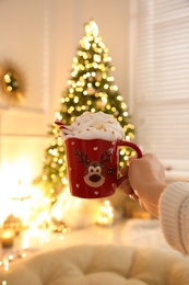 Woman holding cup of delicious drink with whipped cream near Christmas tree indoors, closeup