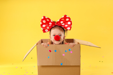 Little girl with large bow and clown nose in cardboard box on yellow background. April fool's day