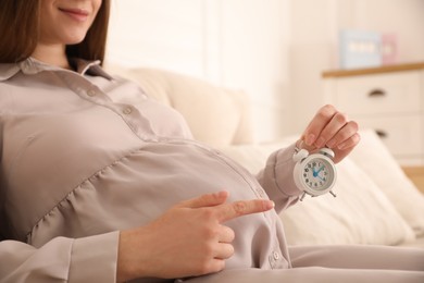 Young pregnant woman pointing at alarm clock near her belly indoors, closeup. Time to give birth
