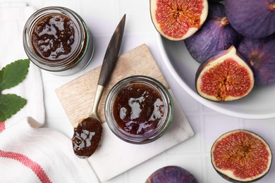 Photo of Glass jars of tasty sweet fig jam and fruits on white tiled table, flat lay