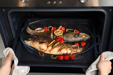 Photo of Woman taking out baking tray with sea bass fish and garnish from oven, closeup