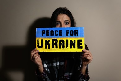 Image of Sad woman holding sign with phrase Peace for Ukraine near beige wall