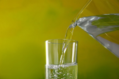 Pouring water from jug into glass on color background, closeup