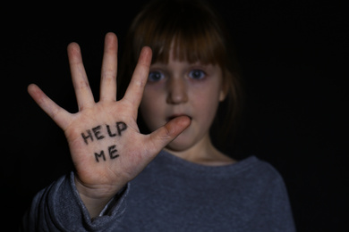 Photo of Abused little girl showing palm with phrase HELP ME on black background, focus on hand. Domestic violence concept
