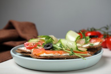 Tasty rye crispbreads with salmon, cream cheese and vegetables served on white wooden table, closeup