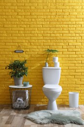 Stylish bathroom with toilet bowl, green plants and decor elements near yellow brick wall. Interior design