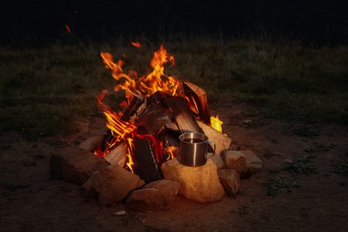 Metal mug with drink near beautiful bonfire outdoors at night. Camping season