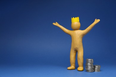 Plasticine figure with crown on head near stacked coins against blue background. Space for text