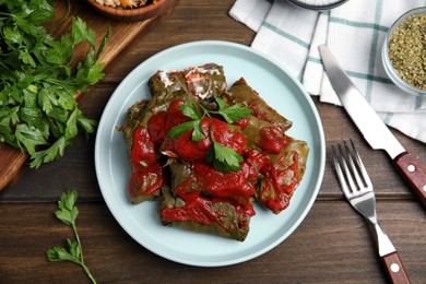 Plate of delicious stuffed grape leaves with tomato sauce, parsley and cutlery on wooden table, flat lay