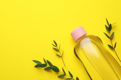 Bottle of baby oil and green leaves on yellow background, flat lay. Space for text