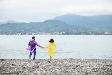 Young couple in raincoats enjoying time together under rain on beach, back view. Space for text