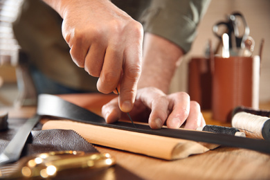 Photo of Man making holes in leather belt with stitching awl at table, closeup
