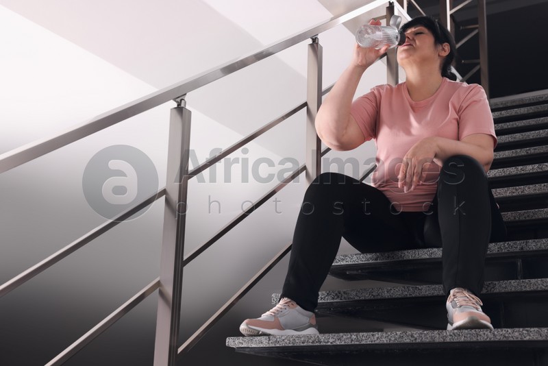 Photo of Tired overweight mature woman drinking water from bottle on stairs indoors. Space for text