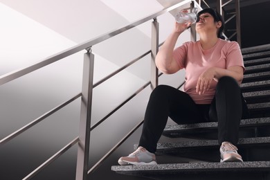 Photo of Tired overweight mature woman drinking water from bottle on stairs indoors. Space for text