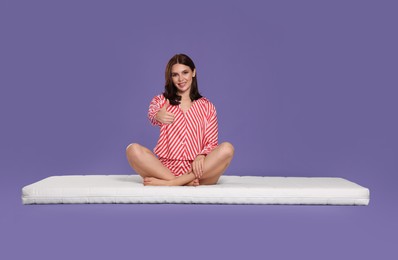 Photo of Young woman sitting on soft mattress and showing thumbs up against light purple background