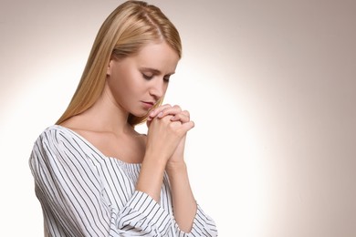 Religious young woman with clasped hands praying against light background. Space for text