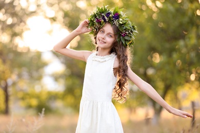 Cute little girl wearing wreath made of beautiful flowers outdoors on sunny day