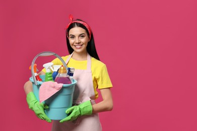 Young housewife holding bucket with cleaning supplies on pink background, space for text