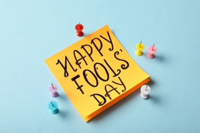 Sticky note with phrase Happy Fools' Day and push pins on light blue background