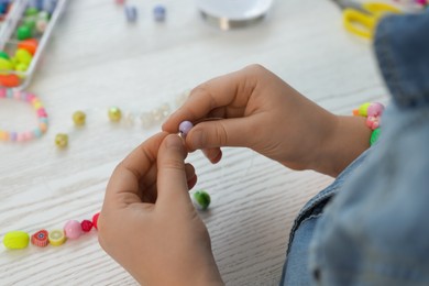 Little girl making beaded jewelry at white wooden table, closeup
