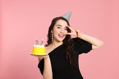 Photo of Coming of age party - 21st birthday. Smiling woman showing peace sign and holding delicious cake with number shaped candles on pink background