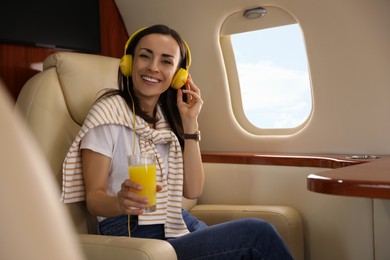 Young woman with glass of juice and headphones listening to music in airplane during flight