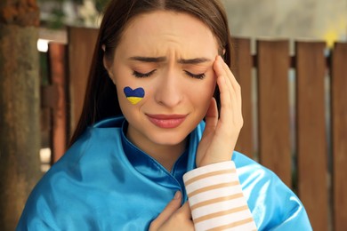 Photo of Sad young woman with drawing of Ukrainian flag on face outdoors, closeup