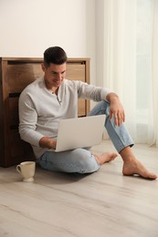 Man with cup of drink and laptop sitting on warm floor at home. Heating system