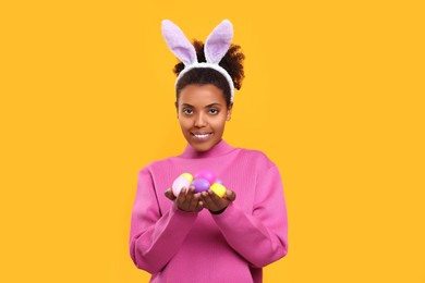 Photo of Happy African American woman in bunny ears headband holding Easter eggs on orange background