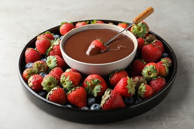 Fondue fork with strawberry in bowl of melted chocolate surrounded by different berries on grey table