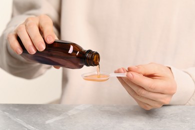 Photo of Woman pouring syrup from bottle into dosing spoon at table, closeup. Cold medicine