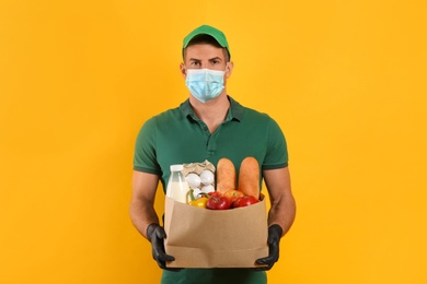 Courier in medical mask holding paper bag with food on yellow background. Delivery service during quarantine due to Covid-19 outbreak