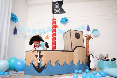 Little boy playing with binoculars in pirate cardboard ship at home. Child's room interior