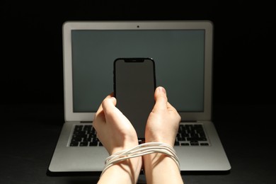 Woman with tied hands holding smartphone near laptop on dark background, closeup. Internet addiction