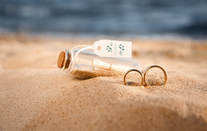 Gold wedding rings and invitation in glass bottle on sandy beach, closeup