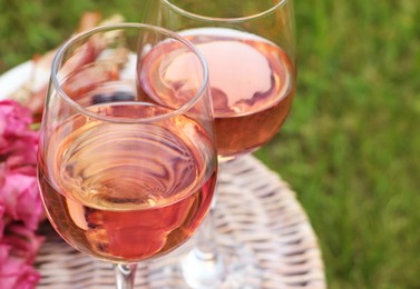 Photo of Glasses of delicious rose wine and flowers on picnic basket outdoors, closeup